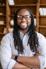 Vertical close-up portrait of cheerful African-American businessman wearing stylish eyeglasses with dreadlocks, a guy stands with arms crossed and looking at camera with a friendly toothy smile