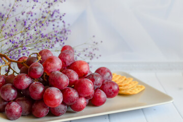 bunch of grapes and cracker on plate. Still life with snack for wine. Red grapes on beige platter on light background. sweet grapes on plate with cheese crackers on white background, space for text