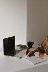 Wall Mural - Minimalist home office workspace with laptop, coffee cup, clipboard. Woman write in a paper sheet notebook. Morning work, business concept. Sunlight shadows on the wall.