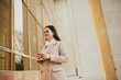 © eduard - Attractive cheerful girl in beige coat and eyeglasses with coffee to go walking outdoor near building.