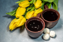 Two Cups Of Tea Or Coffee With Sweets In Coconut Flakes. Tea, Yellow Tulips On A Gray Background. Close-up
