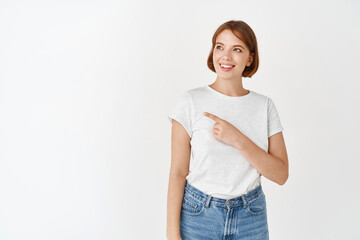 Beautiful natural girl without make-up, white teeth, smiling and pointing left at logo, showing banner, standing in t-shirt on studio background