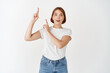 © Cookie Studio - Surprised and excited young girl in t-shirt, poiting fingers up, gasping amazed and smiling, showing cool promo, standing on white background