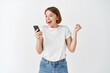 © Cookie Studio - Happy woman winning on smartphone video game, raising hand up and cheering, shouting yes with joy, achieve online goal, standing on white background