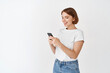 © Cookie Studio - Portrait of smiling woman texting message, reading phone screen. Happy woman chatting on app, standing against white background