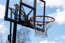 A Portrait Of A Recreational Outdoor Basketball Rim With A Net And A Plexiglass Backboard With Blue Lines On It In Front Of A Blue Sky. The Basketball Hoop Or Ring Is Orange And Its Pole Is Black.