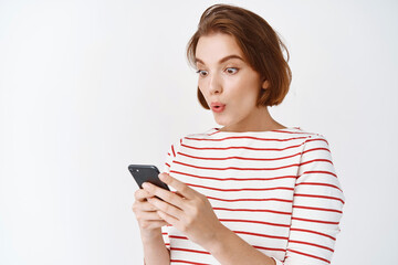 Wow check this out. Surprised and excited girl looking at mobile phone screen, online shopping with big discounts, standing in casual clothes against white background