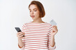 © Cookie Studio - Attractive young woman with short hair, natural light make-up, paying with plastic credit card and smartphone. Girl making order in internet via mobile phone, white background