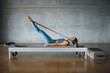 © BASILICOSTUDIO STOCK - Side view of a female Pilates Instructor lying down on a bed of a Pilates Reformer machine, pulling a stretch band with her feet, legs, training in a big Industrial Fitness Gym Studio. Full length.
