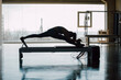 © BASILICOSTUDIO STOCK - Fit female instructor practicing Pilates exercises routine on a Reformer Bed Machine, slim woman working out stretching legs, doing a split, in a big Fitness Gym with big windows and street views.