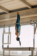 © BASILICOSTUDIO STOCK - Fit woman hanging upside down from a Cadillac Machine, Trapeze Table, Strong pilates instructor doing a straight pose in a big Pilates Studio with wide windows and outside views.