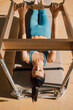 © BASILICOSTUDIO STOCK - High angle view of a woman lying down on a Pilates trapeze table, wearing blue sports clothing, doing an arm exercise routine, in a sunny gym with natural sun light, full length.