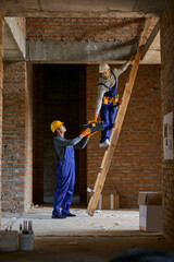 Help. Full length shot of young workman in blue overalls and hard hat giving toolbox to his colleague on ladder while working on house construction