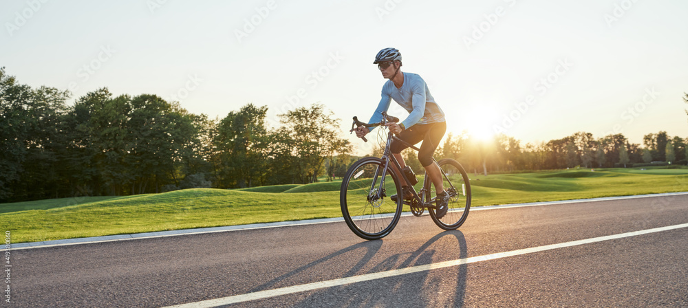 Full length shot of professional male racer in sportswear and helmet training, riding road bike on a cycle path in the park at sunset - obrazy, fototapety, plakaty 