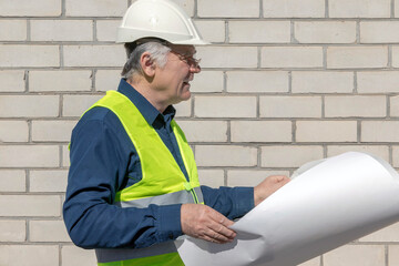 An attentive architect in a white helmet examines a project. On a bright sunny day. Architect with diagrams in hands on a white brick background.