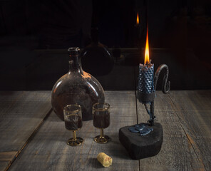 Wall Mural - An old dusty bottle of dark brown bottle with alcohol, two goblet and a metal candlestick holder with a candle on a wooden table on a dark background.