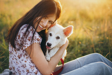 Wall Mural - Happy woman cuddling with cute white puppy in summer meadow in  sunset. Happiness. Summer vacation