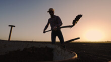 Male Farmer Shovels Earth In Wheelbarrow On Farmland Wearing Straw Hat Plaid Shirt Rubber Boots Sunglasses At Sunset