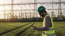 Electrical Engineer Wearing A Helmet And Safety Vest Working With Tablet Near High Voltage Electrical Lines Power Station During Sunset