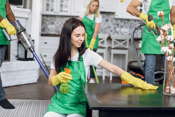 Wall Mural - Attractive happy young brunette woman wiping table with microfiber rag and detergents, while another members of cleaning company vacuuming client's house.