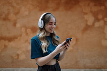Young Focused Happy Female With Wireless Headset Around Neck Text Messaging On Cellphone While Standing Near Wall In Town On Brown Background