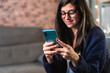 © Alvaro Sanchez/ADDICTIVE STOCK - Positive female freelancer in eyeglasses surfing cellphone and sitting near table and shelves with decorations while working remotely from home
