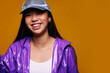 © Victor Torres/ADDICTIVE STOCK - Portrait of happy Asian young woman. She wears a purple jacket and a grey cap and is looking at camera smiling against a yellow background