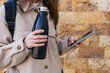 © David Munoz/ADDICTIVE STOCK - Crop anonymous female in warm coat with backpack holding reusable water bottle and browsing modern mobile phone against building wall on street