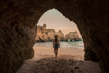 Back View Of Unrecognizable Female In Casual Clothes And Hat Standing On Sand In Entrance Of Cave Near Sea At Sunset In Benagil Caves In Algarve, Portugal