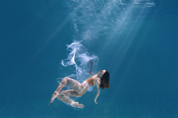 Wall Mural - Fashionable and athletic girl free diver alone in the depths of the ocean. Swimmer brunette diving deep in ocean on blue underwater background. Pollution, plastic and ecology concept