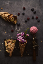 From Above Of Scoop Of Pink Ice Cream Placed On Table With Wafer Cone With Flowers And Scattered Berries On Dark Background