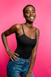 © Luis Manuel Munoz/ADDICTIVE STOCK - Confident young fit African American female model in black top and jeans smiling and looking at camera while standing against pink background with hand on waist