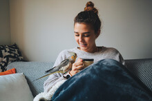 Happy Young Lady In Warm Sweater Smiling And Drinking Hot Coffee While Relaxing On Sofa With Adorable Cockatiel Bird On Hand