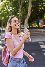 Side View Of Cheerful Young Female With Backpack And Bottle Of Water Standing In Green Park In Sunny Day In Madrid