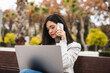 © Luis Tenza/ADDICTIVE STOCK - Concentrated businesswoman sitting on bench with laptop and talking smartphone during remote work in urban park