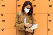 Female Student Wearing Protective Mask Standing With Pile Of Books Near Lockers In University Corridor And Looking At Camera During Coronavirus Epidemic