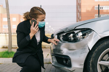 Unrecognizable Young Female Driver In Casual Clothes And Face Mask Talking On Mobile Phone While Standing Near Modern Damaged Car After Accident On City Road