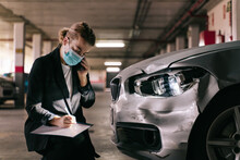 Unrecognizable Concentrated Female Insurance Officer In Formal Suit And Protective Mask Talking On Mobile Phone While Inspecting Car Damages And Filling Out Claim After Accident