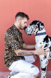 © Sergio Victor Vega/ADDICTIVE STOCK - Side view of young unshaven male in casual clothes and adorable obedient Harlequin Great Dane dog hugging each other against red background