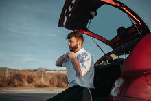 Side View Of Young Bearded Male Driver Sitting In Opened Car Trunk And Taking White Shirt Off Looking Away