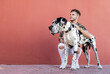 © Sergio Victor Vega/ADDICTIVE STOCK - Young bearded guy in stylish outfit sitting on haunches and stroking obedient Harlequin Great Dane dog against red wall on street