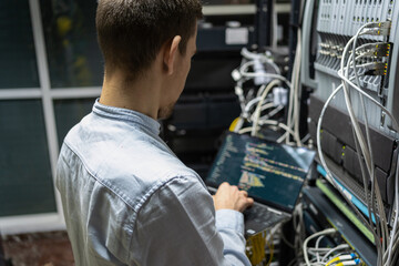 Back view of crop male assistant putting cable in router while using laptop for checking network system