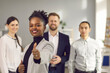 © Studio Romantic - Satisfied dark skinned female client showing thumb up standing against the background of a young startup team. Concept of positive feedback and recommendations. Selective focus.