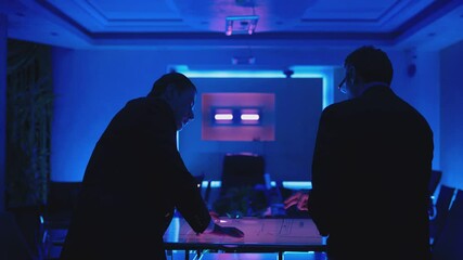 Two men in black suits meeting at dark office room and using a modern, futuristic big touch screen tablet. Businessmen looking at map illuminated on digital table, discussing and talking . Slow Motion
