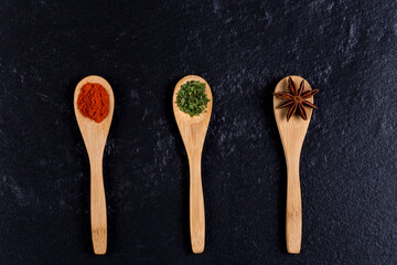 Three wooden spoons with spices on black stone background