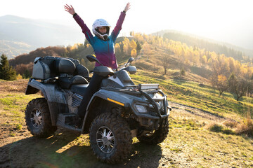 Happy woman driver in protective helmet enjoying extreme riding on ATV quad motorbike in summer mountains at sunset.