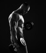 Muscular men bodybuilder is working out in gym, lifting alternately two dumbbells, doing exercises for biceps and looking down over black background. Young man lifting weights. Black and white 