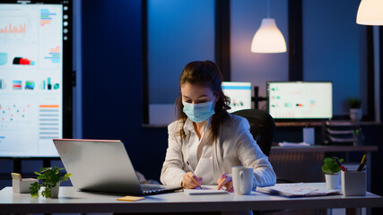 Manager woman with face mask working overtime in new normal business office checking team project, taking notes, analysing financial documents sitting at desk late at night during global pandemic