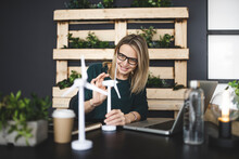 Pretty, Young And Blond Woman With Stylish, Modern Black Glasses Sits In A Sustainable Office And Works With Pinwheel Wind Turbine Models