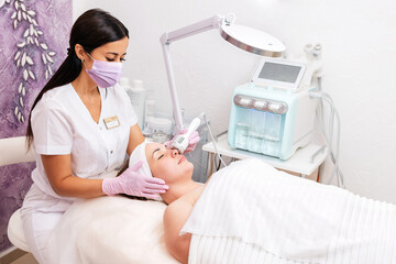 Wall Mural - New normal. A cosmetologist in rubber gloves and medical mask does a rf-lifting procedure on the face. In the background, the Rf cosmetology device. Concept of professional cosmetology and health